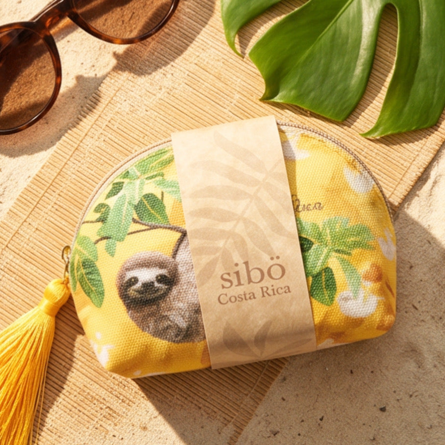Yellow pouch with sloth design and 'sibo Costa Rica' label on a sandy surface with sunglasses and a leaf.